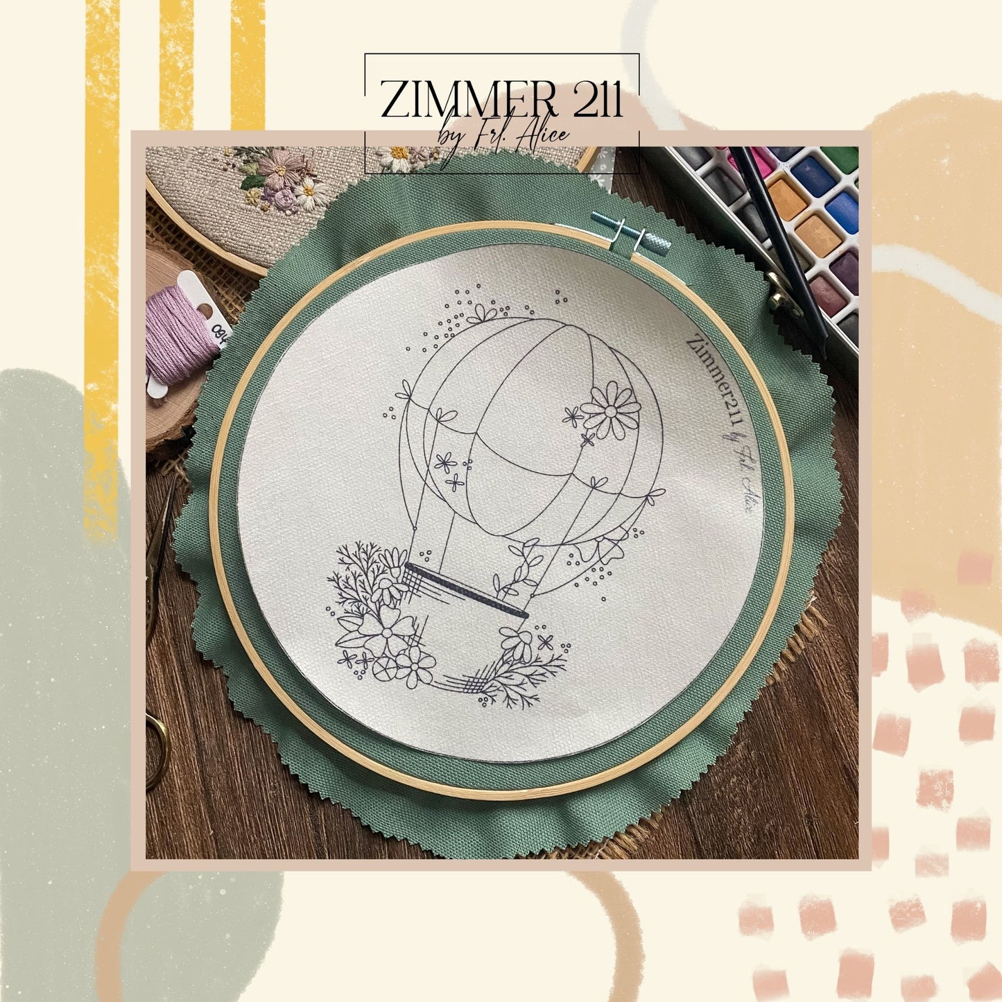 Nahaufnahme einer Stickvorlage von Zimmer211: Ein Heißluftballon mit Blumenmotiven als schwarze Linienzeichnung auf weißem Stoff, eingespannt in einen Holzrahmen mit grünem Stoffuntergrund. Rechts ist der dezente Schriftzug „Zimmer211 by Frl. Alice“ zu sehen. Umgeben von Stickzubehör und einem Aquarellkasten auf dunklem Holz.